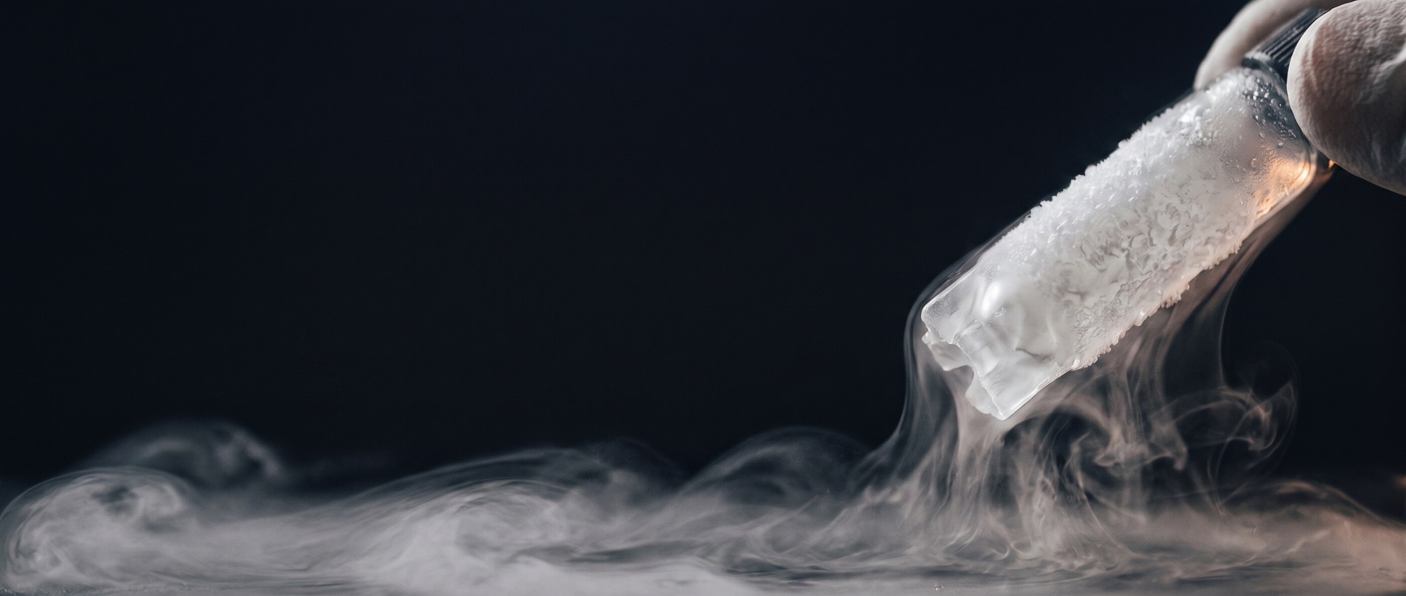 A frost-covered cryovial of cell stock held in tongs, cold vapour from liquid nitrogen drifting below