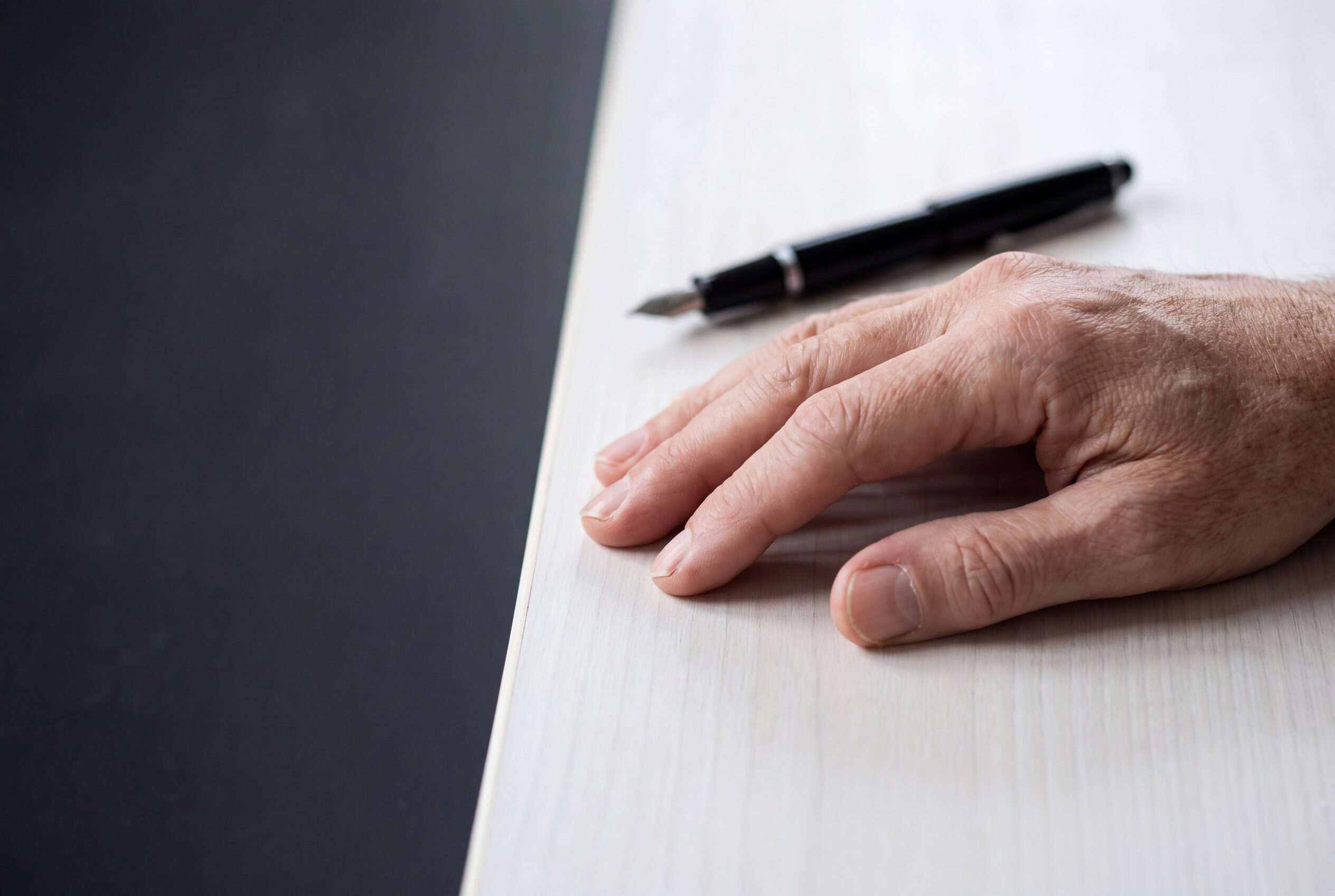 An open hand resting on a table beside a pen just out of reach