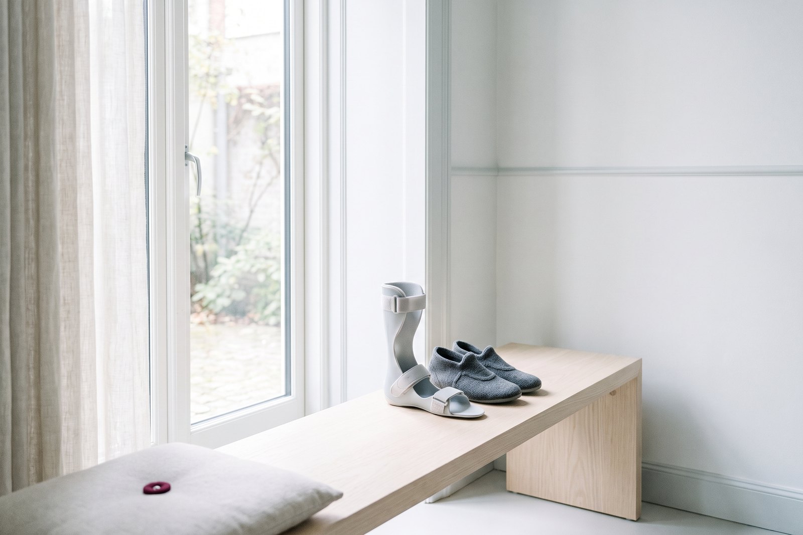 An ankle-foot orthosis and soft shoes on a wooden bench by a window